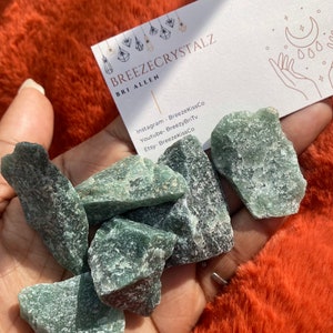 Puede incluir: Un pu&ntilde;ado de cristales verdes &aacute;speros. Se ve una tarjeta de visita con el texto "BREEZECRYSTALZ" y las redes sociales. Los cristales se sostienen sobre un fondo naranja vibrante y difuso.