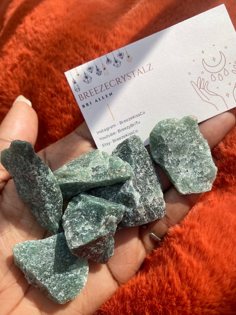 Puede incluir: Un pu&ntilde;ado de cristales verdes &aacute;speros. Se ve una tarjeta de visita con el texto "BREEZECRYSTALZ" y las redes sociales. Los cristales se sostienen sobre un fondo naranja vibrante y difuso.