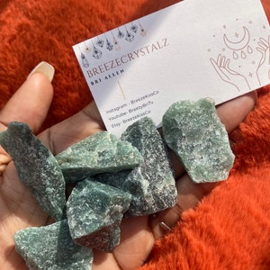 Puede incluir: Un pu&ntilde;ado de cristales de aventurina verde en bruto. Tambi&eacute;n se ve una tarjeta de visita con el texto "BREEZECRYSTALZ". Los cristales se exhiben sobre una superficie naranja vibrante y texturizada.