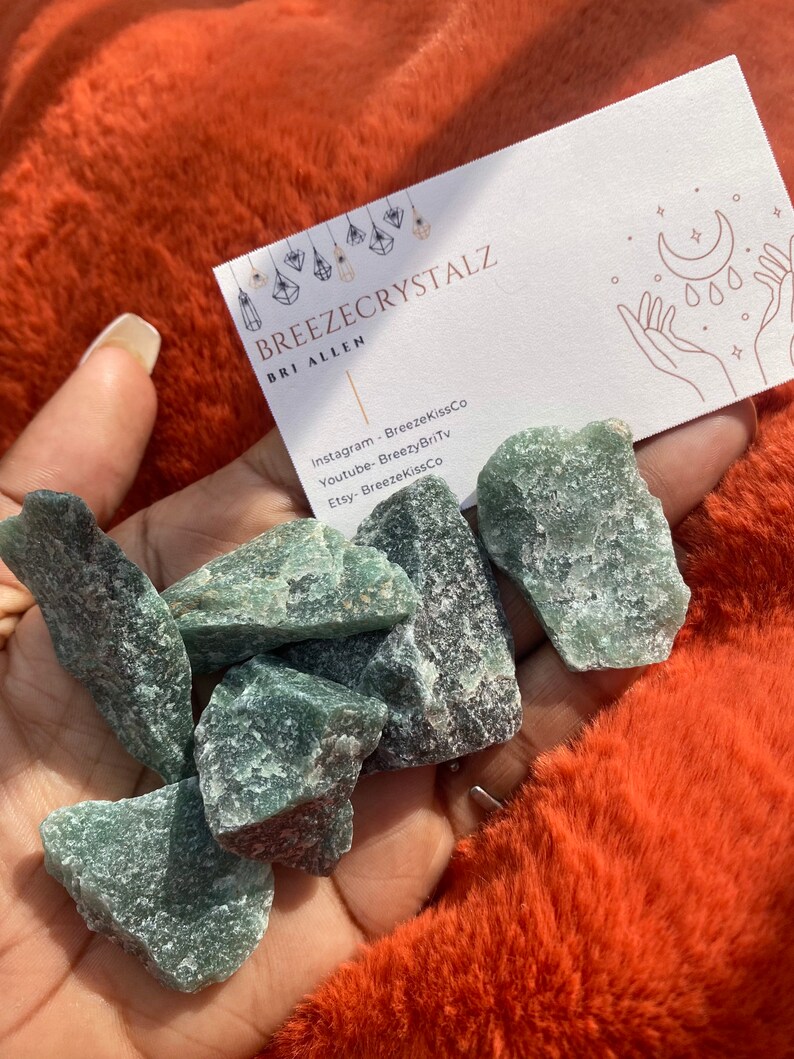 Puede incluir: Un pu&ntilde;ado de cristales de aventurina verde en bruto. Tambi&eacute;n se ve una tarjeta de visita con el texto "BREEZECRYSTALZ". Los cristales se exhiben sobre una superficie naranja vibrante y texturizada.