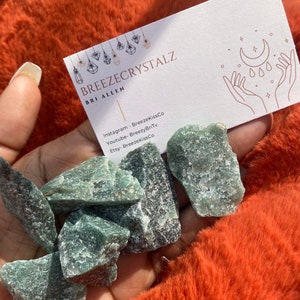 Puede incluir: Una mano sostiene varios cristales de aventurina verde en bruto. Tambi&eacute;n se ve una tarjeta de visita con el texto "Breezecrystalz" y las redes sociales. Los cristales son de color verde natural y tienen una superficie texturizada.