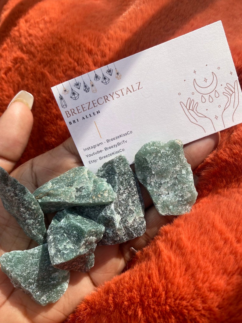 Puede incluir: Una mano sostiene varios cristales de aventurina verde en bruto. Tambi&eacute;n se ve una tarjeta de visita con el texto "Breezecrystalz" y las redes sociales. Los cristales son de color verde natural y tienen una superficie texturizada.