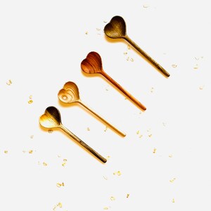 Op de afbeelding: Vier houten hartvormige lepels in verschillende bruintinten, gerangschikt in een rij op een witte achtergrond. De lepels zijn bezaaid met gouden glitter.