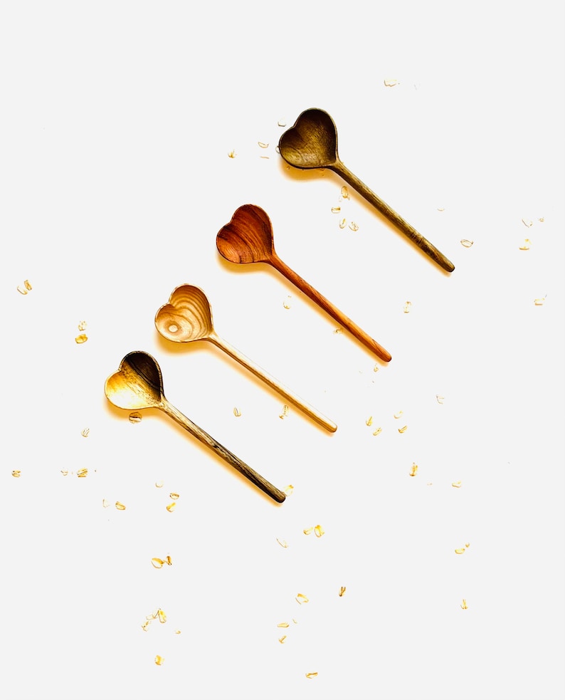 Op de afbeelding: Vier houten hartvormige lepels in verschillende bruintinten, gerangschikt in een rij op een witte achtergrond. De lepels zijn bezaaid met gouden glitter.