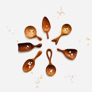 May include: A set of seven wooden spoons with a natural finish. The spoons are arranged in a circle on a white background. Each spoon has a different shape and size. Some of the spoons have a small amount of oats in them.
