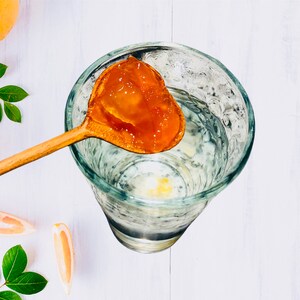 Op de afbeelding: Een houten lepel houdt een schepje oranje gelei boven een glas water. De gelei is doorschijnend en heeft een licht troebele uitstraling.