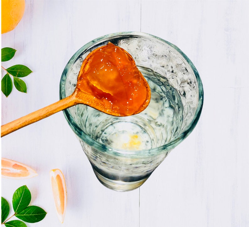 Op de afbeelding: Een houten lepel houdt een schepje oranje gelei boven een glas water. De gelei is doorschijnend en heeft een licht troebele uitstraling.