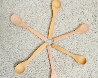 May include: Six wooden spoons arranged in a star shape on a white fluffy surface.