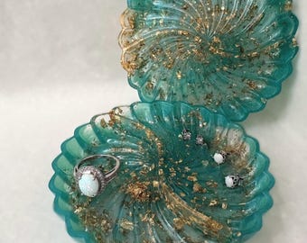 May include: A teal resin trinket dish with a scalloped edge and gold leaf flakes. The dish is open, revealing a matching lid resting behind it. A silver ring with an oval white stone and three small stud earrings are placed inside the dish.