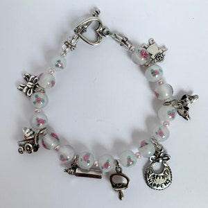 May include: A white beaded bracelet with silver charms. The charms include a baby bottle, a pacifier, a baby carriage, a baby doll, a bow, and a bib that says "Sh*t Happens".