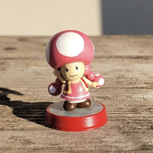 Puede incluir: Figura de un personaje de Super Mario Bros. La figura tiene un gorro de hongo rosa con lunares blancos, un vestido rosa y zapatos marrones. Está sobre una base roja con un círculo gris.