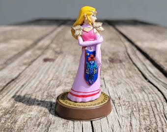 以下が含まれることがあります： 金色の王冠とブロンドの髪をしたピンクと金のプリンセスの置物。彼女は青と金の模様のついたサッシュが付いたピンクのドレスを着ています。置物は茶色の台座の上に立っています。