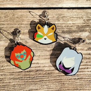 May include: Three colorful keychains featuring cartoon characters. The first keychain is yellow, orange, and green with a fox-like character. The second keychain is green, orange, and red with a character with a large head and a green face. The third keychain is purple, white, and gray with a character with long white hair and a purple dress.
