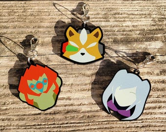 May include: Three colorful keychains featuring cartoon characters. The first keychain is yellow, orange, and green with a fox-like character. The second keychain is green, orange, and red with a character with a large head and a green face. The third keychain is purple, white, and gray with a character with long white hair and a purple dress.