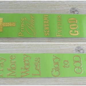 Könnte beinhalten: Zwei grüne Lesezeichen mit goldenen Glitzerbuchstaben. Das obere Lesezeichen hat ein goldenes Kreuz, die Worte "Fürst des Friedens", "Diener", "Vater" und "Gott". Das untere Lesezeichen hat die Worte "Mehr Beten", "Weniger Sorgen", "Ehre sei Gott" und ein goldenes Fischsymbol.