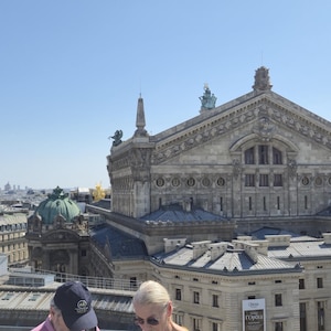 Puede incluir: Vista aérea del Palacio Garnier en París, Francia, bajo un cielo azul claro. El edificio ornamentado presenta detalles de cantería y una gran cúpula. Otros edificios y tejados llenan el fondo.