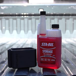 May include: A bottle of STA-BIL Storage Fuel Stabilizer with a black cap and a black rectangular object. The bottle contains a red liquid. The text "STA-BIL STORAGE FUEL STABILIZER AMERICA'S #1" is visible on the label.