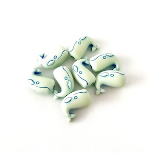 May include: A group of seven light green whale-shaped beads with blue outlines. The beads are arranged in a circle with the tails pointing inwards.