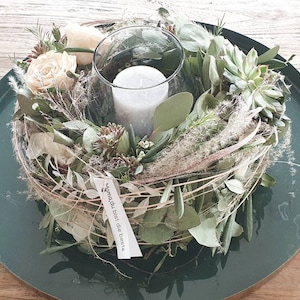 Könnte beinhalten: Ein grünes Tablett mit einer runden Anordnung aus Grünpflanzen, weißen Blumen und einer weißen Kerze in einem Glasgefäß. Die Anordnung umfasst Sukkulenten, Eukalyptus und getrocknete Gräser. Ein kleines weißes Etikett mit dem Text "Du bist der Beste" ist an der Anordnung befestigt.