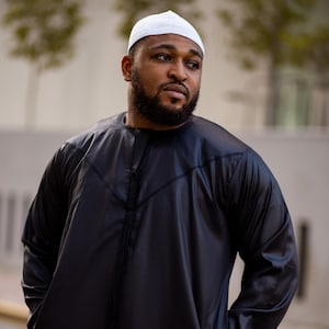 May include: A man wearing a black long-sleeved robe with a white skullcap. The robe has a long, flowing design and a high collar.