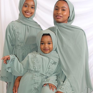 May include: Three individuals wearing matching sage green abayas and hijabs. The abayas feature floral embellishments along the front and sleeves. The individuals are smiling, posing in front of a white backdrop.