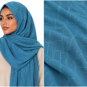 May include: A teal hijab featuring a subtle, tonal cross-hatch or linear textured pattern throughout the fabric. The headscarf is draped around the head and shoulders, showcasing the consistent blue-green color and the intricate, raised stitch details.