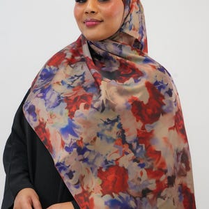 May include: A floral patterned hijab in shades of red, blue, and beige. The hijab is made of a soft, flowing fabric and features a watercolor-like design.