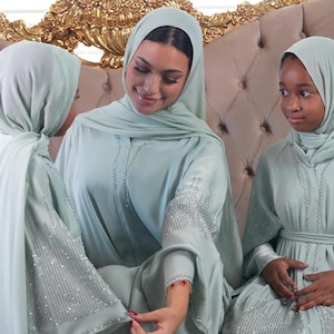 May include: Three women wearing matching light green abayas and hijabs. The abayas have silver embellishments on the sleeves and around the neckline. The women are sitting on a gold and beige ornate sofa.