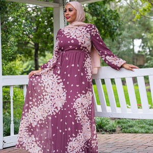 May include: A long-sleeved maxi dress with a floral print in shades of pink and purple. The dress has a pleated skirt and a tie at the waist.