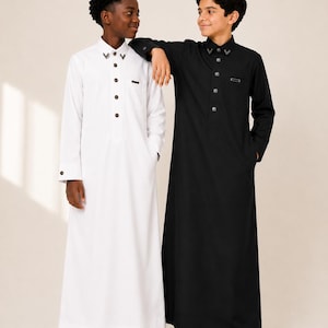May include: Two long-sleeved thobes, one in white and one in black, featuring a structured collar with metallic accents, a front button placket, and a small rectangular chest detail. Both garments reach the ankles and have a straight-cut design.