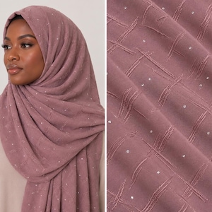 May include: A dusty rose hijab featuring a textured cross-hatch pattern and small, scattered clear rhinestones. The fabric is draped around the head and shoulders, showcasing the intricate embroidered lines and the subtle sparkle of the embellishments.