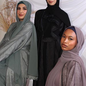 May include: Three women wearing modest attire, including headscarves and flowing dresses. The dresses are in shades of gray, black, and mauve, with intricate patterns and embellishments. The background is a neutral white.