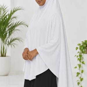 Può includere: Un hijab bianco a tinta unita con una tunica a maniche lunghe abbinata caratterizzata da una silhouette ampia e fluida. Il capo è abbinato a una gonna nera semplice, creando un look a strati con linee pulite e un design coprente.