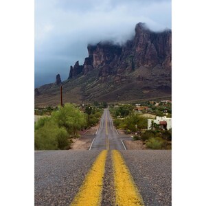 May include: A long, paved road with yellow lines leads towards a mountain range in the distance. The mountains are covered in clouds and mist, creating a dramatic and atmospheric scene.