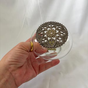 May include: A clear glass bowl featuring a circular, intricate metal lid with a detailed filigree pattern. The lid is primarily silver-toned with a small, central gold-toned accent, showcasing a symmetrical, floral-inspired design that covers the top.