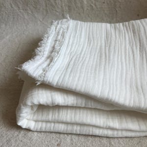 May include: Folded white fabric with a textured, crinkled appearance. The edges of the fabric have a fringed detail. The fabric is layered, showing the folds and texture of the material. The background is a neutral, light-colored surface.