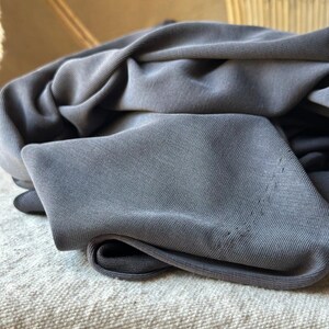 May include: Close-up of a folded, gray fabric. The fabric appears to be a soft knit material, with visible texture and slight variations in color. The folds create shadows and depth, highlighting the fabric's drape.