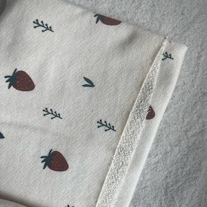 May include: Close-up of a cream-colored fabric with a repeating pattern of red strawberries and green sprigs. The edge of the fabric is trimmed with a textured, off-white border. The fabric is likely part of a garment or textile.