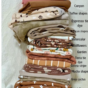 Pode incluir: Uma pilha de tecidos dobrados em várias cores e padrões. As cores incluem rosa bebê, marrom, bege e tons de verde. Os padrões incluem formas, tie-dye, listras e motivos florais. O tecido é rotulado com nomes como "Toffee shapes" e "Desert Canyon."