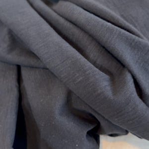 May include: Close-up of dark grey fabric with visible texture and folds. The material appears soft and drapes naturally, suggesting it could be used for clothing or home textiles. The lighting highlights the subtle variations in the fabric's surface.