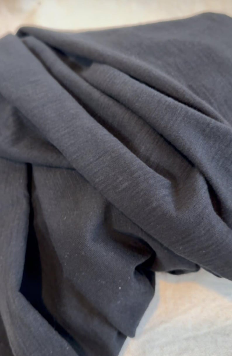May include: Close-up of dark grey fabric with visible texture and folds. The material appears soft and drapes naturally, suggesting it could be used for clothing or home textiles. The lighting highlights the subtle variations in the fabric's surface.