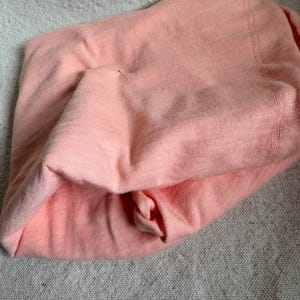 May include: A folded peach-coloured knit fabric, possibly a shirt or top, with a textured appearance. The fabric is soft and appears to be made of a lightweight material. The item is laying on a grey textured surface.