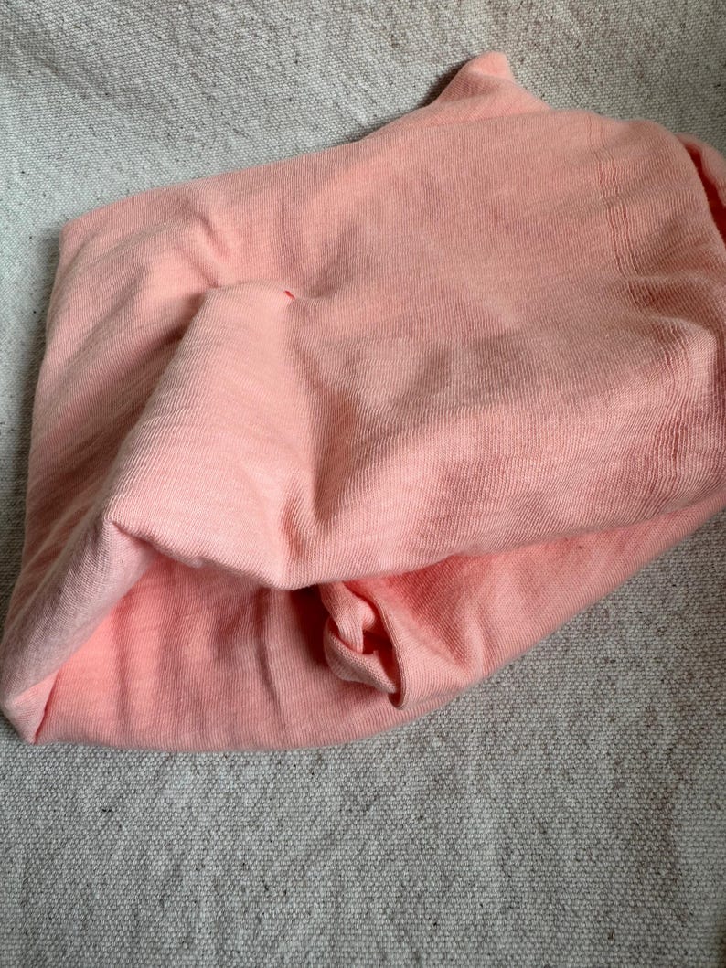 May include: A folded peach-coloured knit fabric, possibly a shirt or top, with a textured appearance. The fabric is soft and appears to be made of a lightweight material. The item is laying on a grey textured surface.
