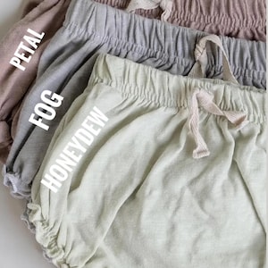 May include: Close-up of four pairs of baby bloomers in Petal, Fog, and Honeydew colours. Each pair has a drawstring waistband and ruffled leg openings. The text "Petal", "Fog", and "Honeydew" are printed on the bloomers.