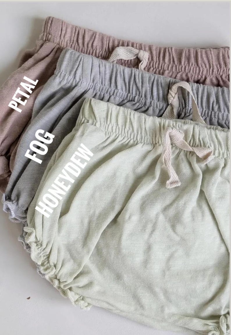 May include: Close-up of four pairs of baby bloomers in Petal, Fog, and Honeydew colours. Each pair has a drawstring waistband and ruffled leg openings. The text "Petal", "Fog", and "Honeydew" are printed on the bloomers.