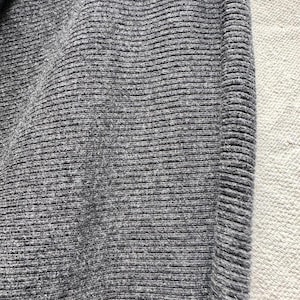 May include: A dark gray knit fabric featuring a consistent horizontal ribbed texture. The material displays a dense, woven pattern with subtle variations in the charcoal gray yarn, creating a soft, tactile surface with a clear vertical seam.