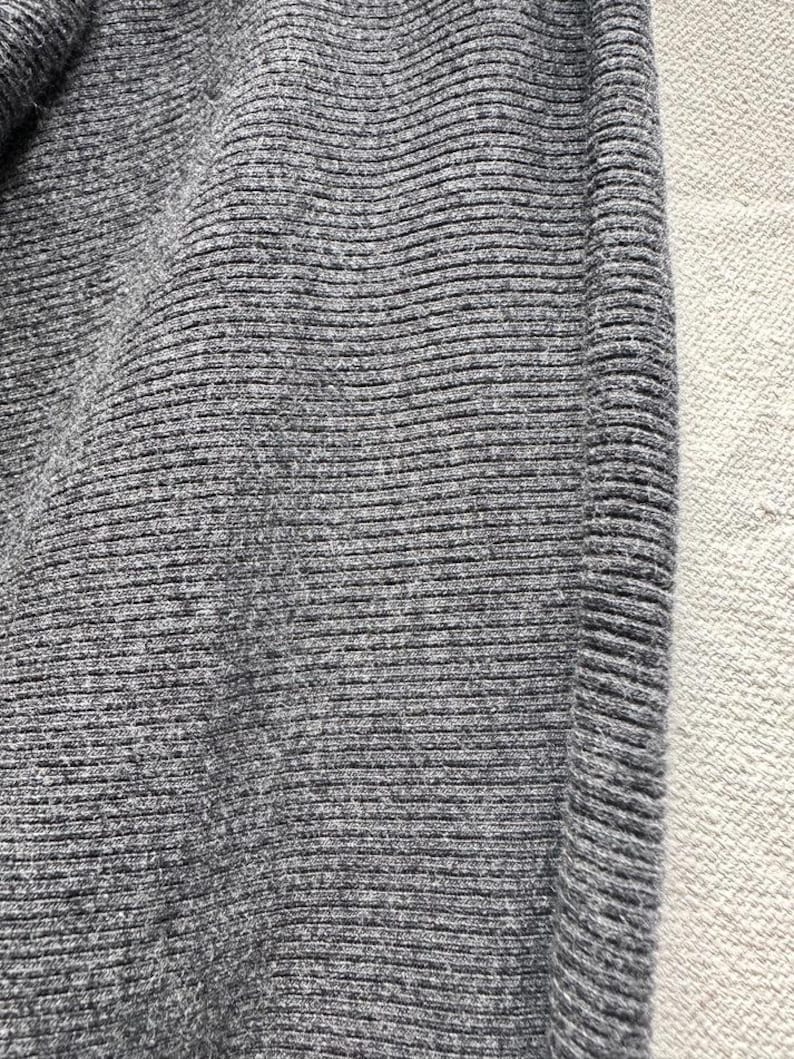 May include: A dark gray knit fabric featuring a consistent horizontal ribbed texture. The material displays a dense, woven pattern with subtle variations in the charcoal gray yarn, creating a soft, tactile surface with a clear vertical seam.