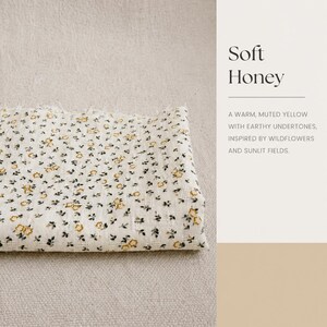 May include: A folded piece of off-white fabric featuring a small-scale floral pattern with yellow blossoms and dark green leaves. The text on the right side reads Soft Honey, A WARM, MUTED YELLOW WITH EARTHY UNDERTONES, INSPIRED BY WILDFLOWERS AND SUNLIT FIELDS.