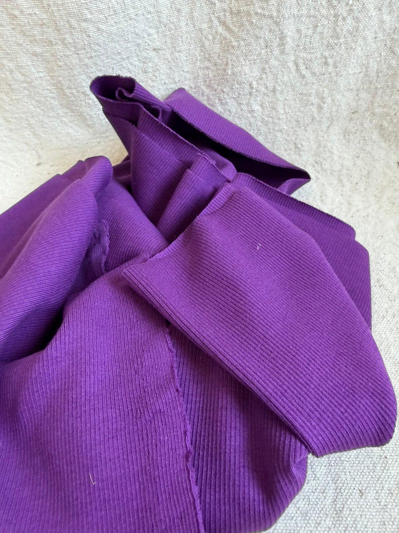 May include: A vibrant purple long-sleeve shirt made of a ribbed knit fabric. The garment features a high neckline and visible vertical ribbing throughout the material, with raw, unfinished edges at the cuffs and collar area, showing a textured surface.