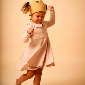 May include: A young girl wearing a pink dress and a gold crown is smiling and twirling.